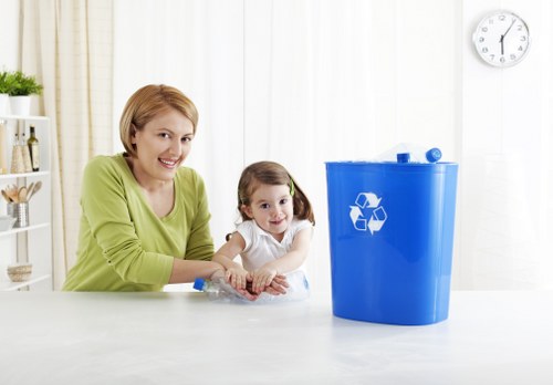 A woman and a young girl with light brown hair are sitting at a white kitchen countertop in a bright, modern kitchen with white cabinets and a window with cream curtains in the background. The woman, wearing a lime-green top, is smiling while holding a clear plastic bottle, and beside her, the girl in a white shirt is also smiling and appears engaged. On the right side of the image, there is a large blue recycling bin with a white recycling symbol on it, placed on the countertop, suggesting waste or rubbish collection. The environment is clean and well-lit, with a simple, functional aesthetic. A wall clock with a white face and black hands is visible on the right side of the background, indicating the time. This setting highlights responsible waste management practices, aligning with services provided by Commercial Waste Removal Kingston, which specialises in rubbish collection and disposal in the local area.