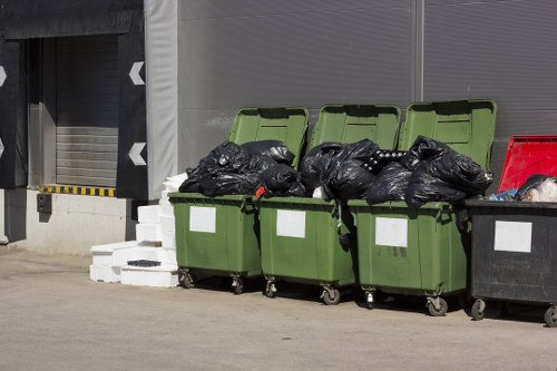 Four large wheeled rubbish bins are positioned in a row outside a commercial building, with three of them having their green lids open and filled with black waste bags. The black waste bags appear tightly packed and are stacked on top of each other within the bins. To the right, a single red bin is visible, partially obscured by the black waste bags, with its lid closed. The bins are placed on a paved surface, and in the background, there is a building wall with metallic and industrial features, including a grey shutter door on the left side. The overall scene suggests a waste collection area, likely associated with commercial waste management services offered by Commercial Waste Removal Kingston, serving areas near Kingston upon Thames or London postcode KT1. The environment appears clean, organized, and typical for rubbish disposal at a commercial or industrial premise, emphasizing the company's role in local waste removal solutions.