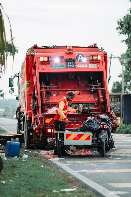 A red metal skip with a weathered, slightly rusty surface is positioned against a wall on a paved urban street. The skip is rectangular with vertical ridges running along its sides, and its paint appe