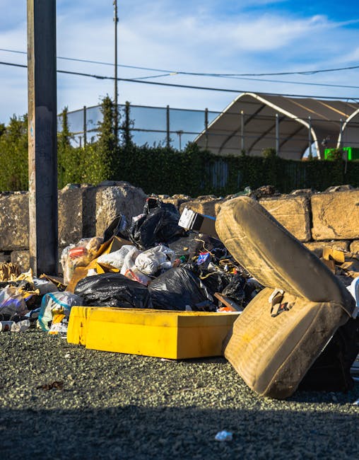 A pile of discarded household items and debris situated outside against a brick wall and concrete pavement. The foreground features a wooden garden trellis with horizontal slats leaning on the ground,